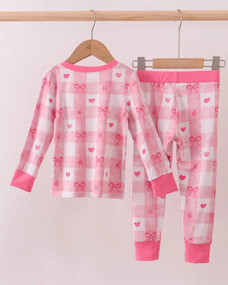 Children's pink and white checkered pajama set with bows and hearts on a hanger against a light pink background.