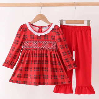 A toddler girl's two-piece clothing set hanging on wooden hangers against a plain white wall. The set includes a red long-sleeve dress with a black plaid pattern, a smocked chest panel, and a white lace collar, paired with matching red pants featuring ruffled hems.