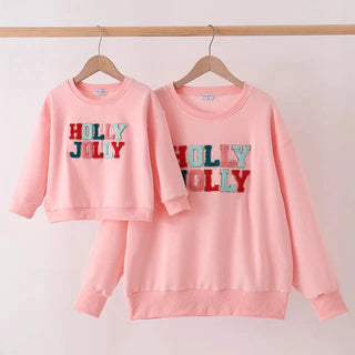 A matching mother and daughter pink sweatshirt set hanging on wooden hangers against a light pink background. The larger adult sweatshirt features 'HOLLY JOLLY' text in red and blue glittery letters, and the smaller child sweatshirt features a smaller version of the same text.