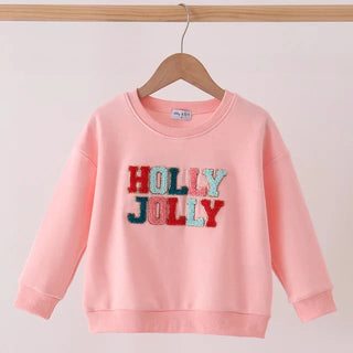 A light pink long-sleeve sweatshirt hanging on a wooden hanger, featuring the words 'HOLLY JOLLY' in red and blue sequins across the chest.