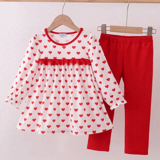 Children's outfit with heart-patterned dress and red pants on hangers against a white background