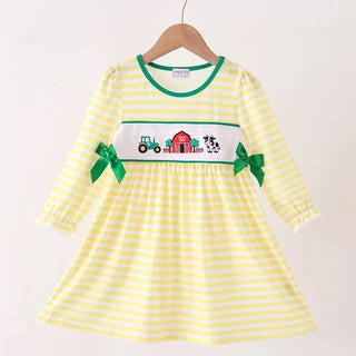 A yellow and white striped long-sleeve baby dress hanging on a wooden hanger. The dress features a white smocked chest panel with a colorful tractor and barn design, green trim, and green ribbon bows on the sleeves.