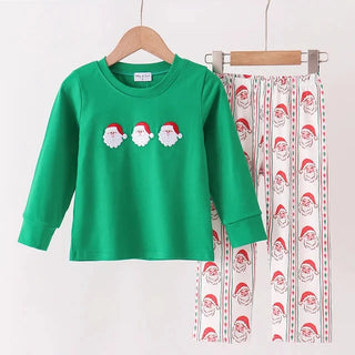 A children's holiday pants set hanging on a wooden hanger bar against a plain white wall. The set includes a bright green long-sleeve top with three Santa Claus faces on the chest, and matching white pants with a vertical pattern of Santa Claus faces and red hats.