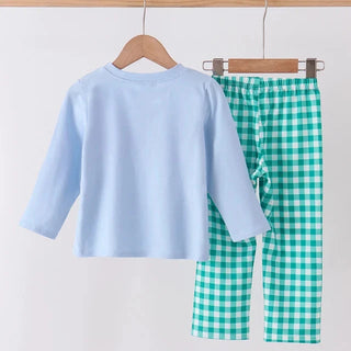 A children's two-piece clothing set hanging on wooden hangers from a wooden rod. The set includes a light blue long-sleeve top and green and white gingham check pants.