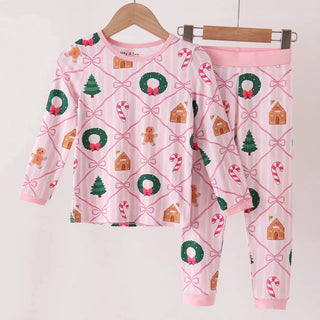 A two-piece children's pajama set hanging on wooden hangers against a plain light background. The set includes a long-sleeve top and matching pants in light pink with a Christmas-themed print featuring green wreaths, candy canes, gingerbread houses, and small trees.