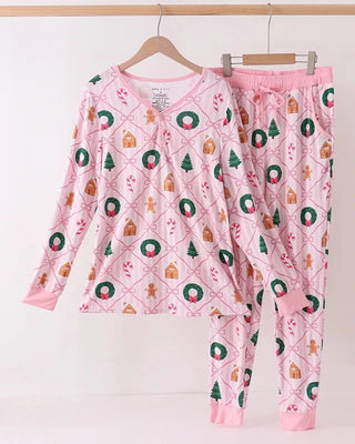 A two-piece women’s pajama set hanging on wooden hangers against a white wall. The set includes a long-sleeve top and matching pants in light pink, decorated with a holiday-themed print featuring gingerbread houses, Christmas trees, wreaths, and candy canes.