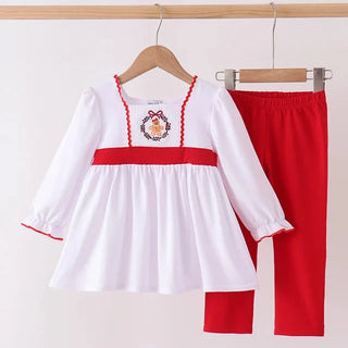 A two-piece toddler girl's outfit set consisting of a white long-sleeve dress with red trim and a red waistband, featuring a small crest embroidery on the chest, paired with solid red pants. The set is displayed on wooden hangers hanging from a wooden clothing rack against a plain off-white wall.