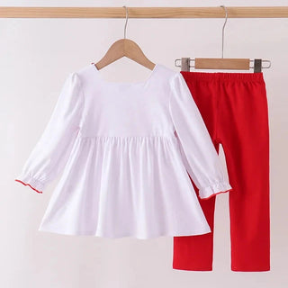 A toddler girl's two-piece clothing set hanging on wooden hangers against a plain light background. The set includes a white long-sleeve smocked blouse with red piping on the sleeve cuffs and solid red pants.