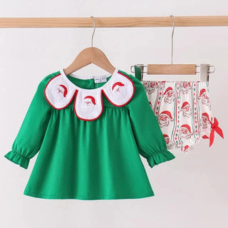 A two-piece toddler Christmas outfit hanging on wooden hangers against a plain white wall. The set includes a bright green long-sleeve top with three white Peter Pan-style collar patches featuring red trim and Santa Claus faces, paired with white shorts printed with a red Santa Claus pattern and a small red bow.