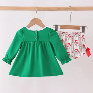 A toddler girl's long-sleeve green dress with puff sleeves and a buttoned neckline, hanging on a wooden hanger next to matching printed shorts featuring Santa Claus illustrations.