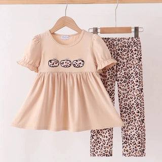A two-piece toddler girl's outfit set hanging on wooden hangers against a plain white wall. The set includes a short-sleeve peplum-style top in light peach-beige with three embroidered pumpkin motifs on the chest, and matching leopard-print leggings in brown, beige, and black.