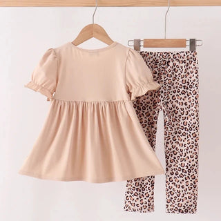 A toddler girl's two-piece outfit hanging on wooden hangers from a clothing rack. The set includes a short-sleeve peplum top in a solid light peach-beige color and matching leopard-print pants in beige, brown, and black.