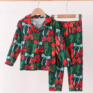 A two-piece children's pajama set featuring a long-sleeve button-up top and matching pants, both printed with a festive red bow and green leaf pattern on a dark green background, hanging on wooden hangers against a plain white wall.