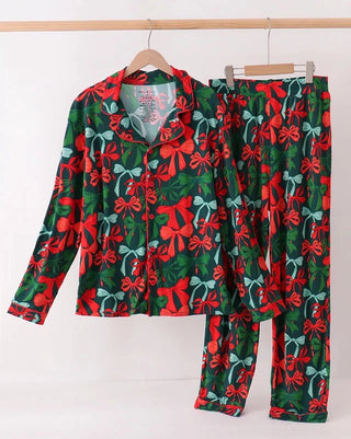 A two-piece pajama set hanging on wooden hangers against a plain white wall. The set includes a long-sleeve button-front top and matching pants, both featuring a festive Christmas pattern of red bows, green pine trees, and red berries on a dark green background.