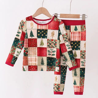 A two-piece children's pajama set hanging on wooden hangers from a light wood rod. The set includes a long-sleeve top and matching pants in a Christmas-themed patchwork pattern with red, green, beige, and white blocks, featuring pine trees, ornaments, and plaid details.