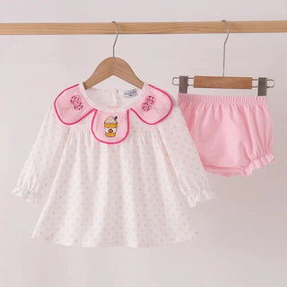 A baby girl's two-piece outfit hanging on wooden hangers against a plain white wall. The set includes a white long-sleeve top with tiny pink polka dots, a pink collar trimmed in pink, and a small embroidered character on the center of the collar, paired with matching pink ruffled bloomers.