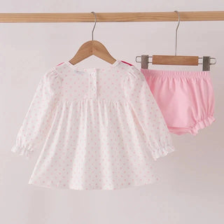 A baby girl's two-piece outfit set hanging on wooden hangers from a wooden rod. The set includes a white long-sleeve top with tiny pink polka dots and a matching pink bloomers set.