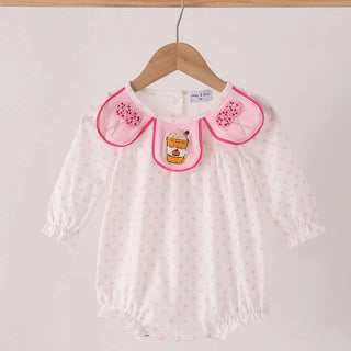A white long-sleeve baby romper with tiny pink polka dots, featuring a pink-trimmed bib collar with embroidered pink strawberries and a central cartoon bear patch, hanging on a wooden hanger.