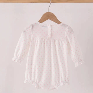 A white long-sleeve baby romper with a tiny pink polka dot pattern, hanging on a wooden hanger from a light wood clothing rack against a plain off-white background.