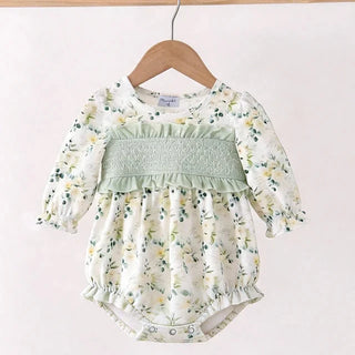 A baby long-sleeve romper with a white and green floral print, featuring a smocked chest panel and ruffled trim, hanging on a wooden hanger against a light pink background.