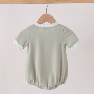 A short-sleeve baby romper in sage green, hanging on a wooden hanger from a light wood clothing rack against a pale pink background.