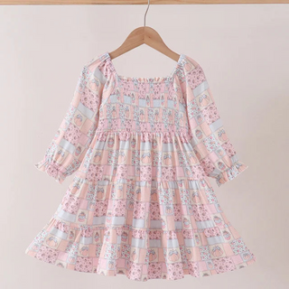 A pink long-sleeve baby dress with a smocked bodice and tiered skirt, featuring a pastel patchwork print with blue, yellow, and white motifs, hanging on a wooden hanger against a light beige wall.