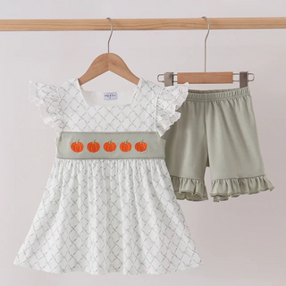 A girl's two-piece outfit set hanging on wooden hangers against a light wall. The set includes a white dress with a subtle geometric pattern, ruffled sleeves, and a light sage green waistband decorated with five orange embroidered pumpkins, paired with matching sage green ruffled shorts.