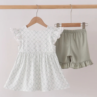 A toddler girl's two-piece outfit hanging on wooden hangers from a light wood rod. The set includes a white smocked dress with a subtle gray trellis pattern and flutter sleeves, paired with sage green ruffle-trimmed shorts.