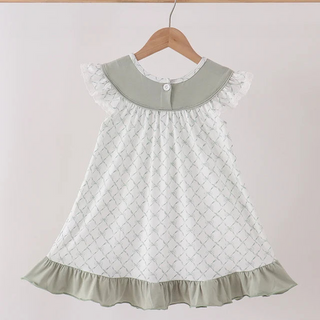 A white girl's dress with a light sage green collar and ruffled trim, hanging on a wooden hanger against a light pink background.