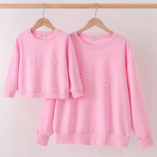 A pink matching mother-and-child sweatshirt set hanging on wooden hangers against a light pink wall. The set includes a small long-sleeve top and a larger long-sleeve top, both with white bow embroidery on the chest.
