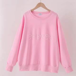 A light pink crewneck sweatshirt with a white embroidered bow pattern across the chest, hanging on a wooden hanger against a pale pink wall.