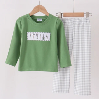 A children's two-piece clothing set hanging on wooden hangers against a plain light background. The set includes a sage green long-sleeve top with a white rectangular chest patch showing kitchen utensils, and matching white and light gray gingham pants.