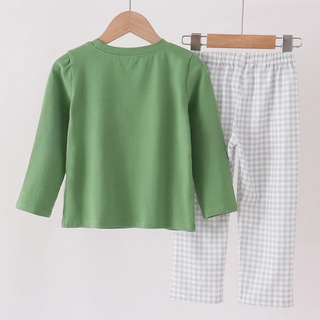 A children's two-piece clothing set hanging on wooden hangers from a wooden rod. The set includes a sage green long-sleeve top and light gray and white gingham check pants.
