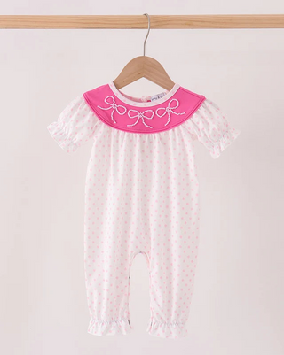 A baby girl's short-sleeve romper in white with tiny pink polka dots, featuring a contrasting bright pink collar with white embroidered bows, hanging on a wooden hanger.