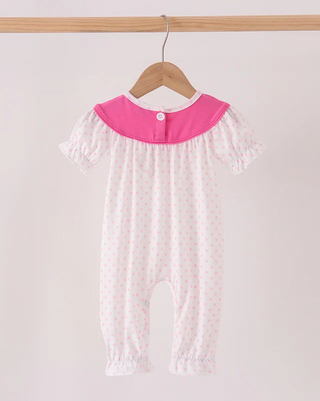 A baby short-sleeve footless romper hanging on a wooden hanger from a wooden rod. The romper is white with tiny pink polka dots and features a contrasting bright pink collar with a single button closure.