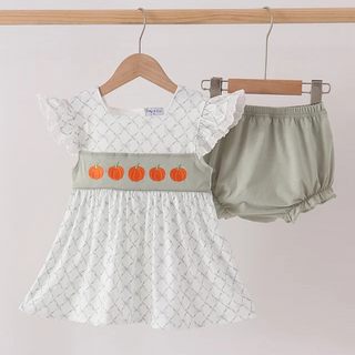 A baby girl's two-piece outfit hanging on wooden hangers against a light pink wall. The set includes a white short-sleeve dress with a subtle gray diamond pattern, a light sage green waistband decorated with five orange pumpkins, and matching sage green bloomers.