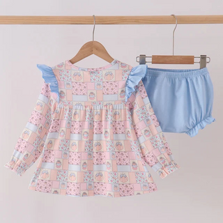 A toddler girl's two-piece outfit set hanging on wooden hangers against a light pink background. The set includes a long-sleeve ruffle-shoulder top in a pastel patchwork print of pink, blue, and white, paired with light blue ruffle-trimmed bloomers.