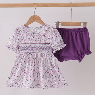 A toddler girl's two-piece outfit set hanging on wooden hangers against a plain light background. The set includes a short-sleeve smocked dress in a white fabric with a purple floral print, and a pair of solid purple ruffled bloomers.