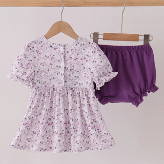A two-piece girls' outfit hanging on wooden hangers against a plain light background. The set includes a short-sleeve white dress with a purple and pink floral print and matching purple bloomers.