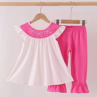 A toddler girl's two-piece outfit hanging on wooden hangers against a light pink wall. The set includes a white short-sleeve top with tiny pink polka dots, a bright pink collar with white embroidered bows, and matching bright pink leggings with ruffled cuffs.