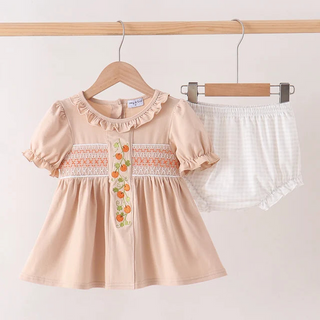 A baby girl's two-piece outfit set consisting of a light peach short-sleeve dress with ruffles, smocked details, and a floral embroidered placket, paired with white gingham check bloomers, hanging on wooden hangers from a wooden clothes rack.