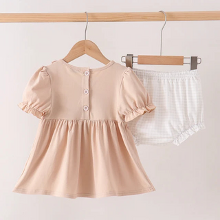 A baby girl's two-piece outfit hanging on wooden hangers against a light beige wall. The set includes a short-sleeve peplum top in a soft peach-beige color with three decorative buttons on the back, and matching white gingham check bloomers with ruffled trim.