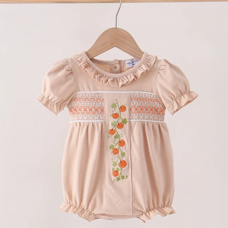 A baby girl's short-sleeve romper in a soft peach-beige color, featuring ruffled trim at the collar, smocked detailing across the chest, and a vertical embroidered floral motif with orange flowers and green leaves. The romper is shown hanging on a wooden hanger from a light wood bar against a plain off-white background.