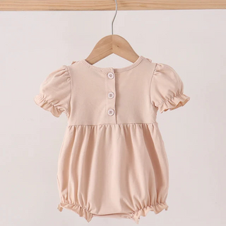 A baby girl's short-sleeve romper in a soft, pale peach-pink color, hanging on a wooden hanger from a wooden clothes rack. The romper features a buttoned neckline with three white buttons and a gathered waist.