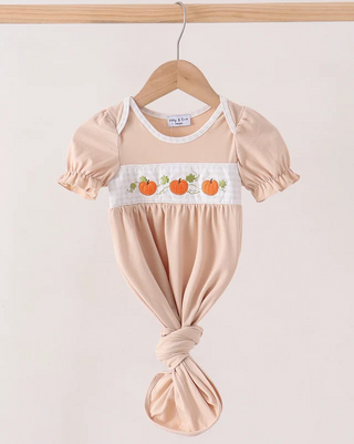 A baby gown in a soft peach-beige color, hanging on a wooden hanger against a light beige background. The romper features short puff sleeves, a white smocked chest panel with embroidered orange pumpkins, and a knotted leg detail.