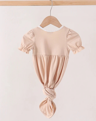 A light peach-pink baby gown with short puff sleeves and a knotted waist, hanging on a wooden hanger against a plain off-white background.