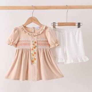 A toddler girl's two-piece outfit set hanging on wooden hangers against a plain light background. The set includes a short-sleeve peach dress with ruffles, a smocked orange and white chest panel with a floral embroidered detail, and white textured shorts with a ruffled hem.