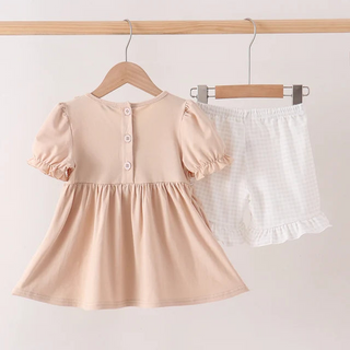 A two-piece toddler girl's outfit set hanging on wooden hangers against a light beige wall. The set includes a short-sleeve peplum dress in a soft peach-beige color with three front buttons, and a pair of white gingham check shorts with a ruffled hem.