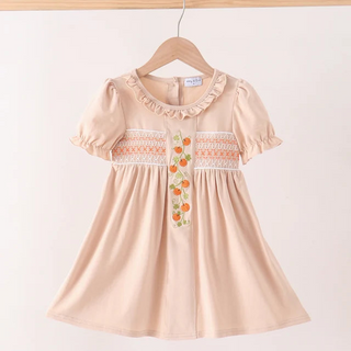A short-sleeve dress in a soft peach-beige color, hanging on a wooden hanger against a plain off-white background. The dress features ruffled trim around the round neckline, white smocked detailing across the chest, and a vertical embroidered floral motif in orange, green, and yellow.