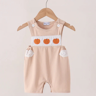 A baby romper in a light peach-beige color, hanging on a wooden hanger. The romper features a white chest panel with three orange embroidered pumpkins and white button details on the shoulder straps and side pockets.